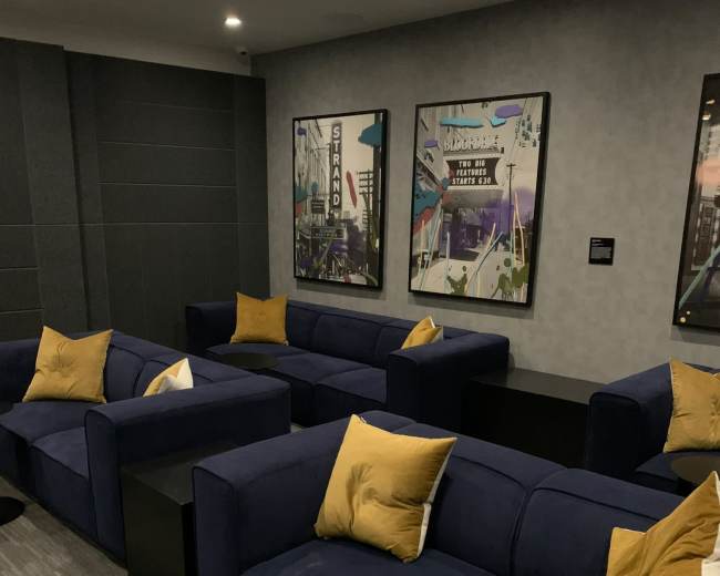The image shows a modern lounge area with navy blue couches, yellow accent pillows, and two framed artworks on the wall, set against a gray backdrop.