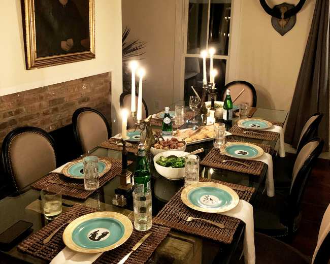 A dining table is set with blue plates, glasses of water, and candles, surrounded by a warm, inviting room featuring a large painting and decorative horns on the wall.