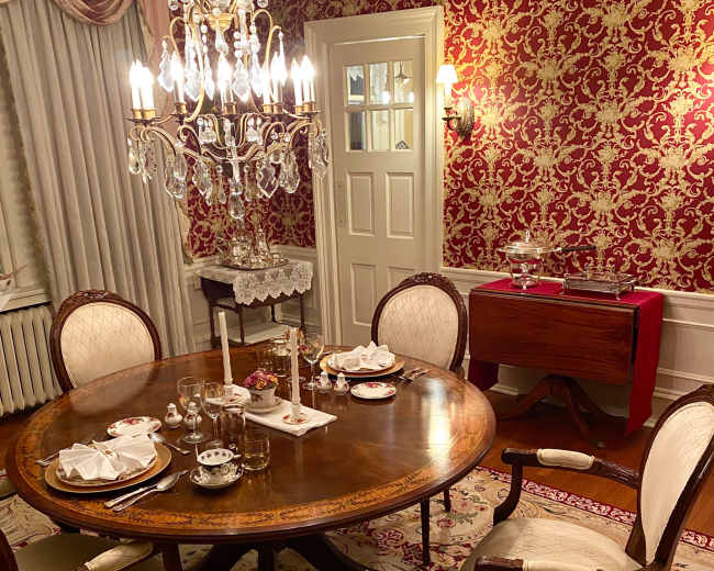 A formal dining room features an ornate round table set with dishes and glasses, surrounded by elegant chairs, with a crystal chandelier hanging above and floral wallpaper covering the walls.
