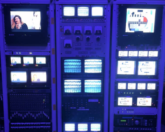 The image shows a large, multi-section electronic display rack filled with various video monitors, control panels, and equipment, illuminated by blue lighting.