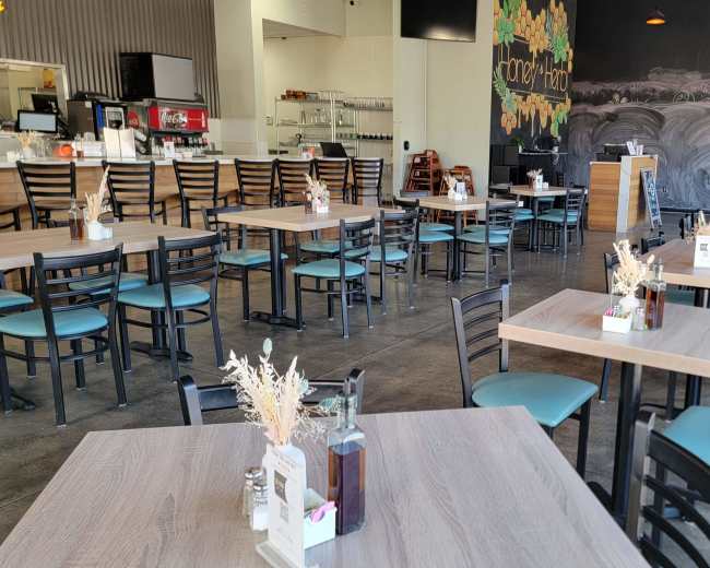 The image shows a spacious, modern restaurant with wooden tables and chairs arranged for dining, and a chalkboard wall in the background.