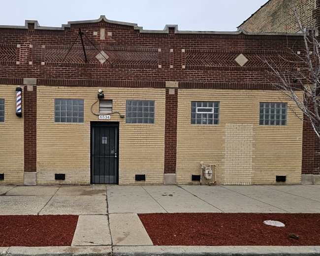 The image shows a single-story brick building with a yellow and red exterior, featuring a black door and several small windows.