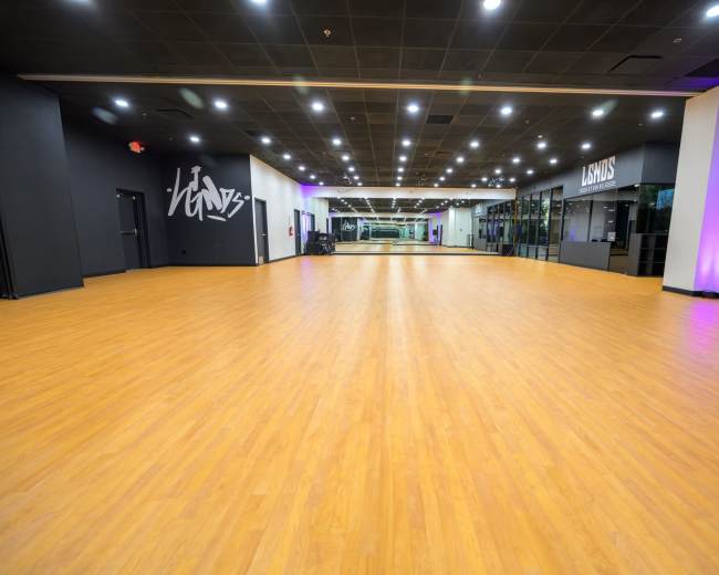 The image shows a spacious dance studio with wooden flooring, brightly lit by overhead lights and featuring mirrors along the walls.