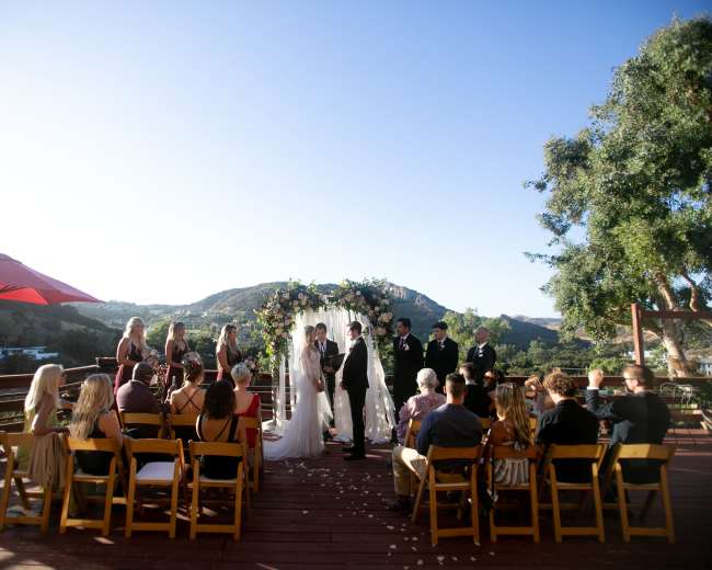An outdoor wedding ceremony takes place on a wooden deck with guests seated in rows, overlooking a scenic landscape.