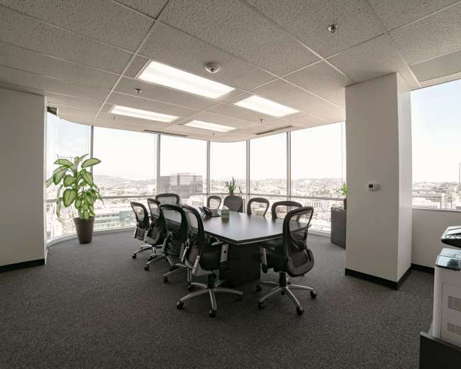 A modern conference room with a large table surrounded by office chairs, featuring floor-to-ceiling windows that overlook a cityscape.