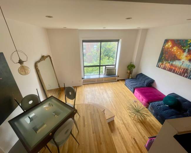 A spacious, well-lit room features a large window, a dark blue couch with a pink section, a round wooden coffee table, and a dining table with transparent chairs.