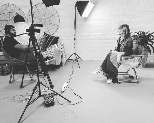 A person sits in a chair during an interview while another person operates a camera on a tripod in a well-lit room with a couch and decorative items.