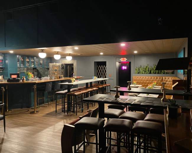 The image shows a dimly lit bar area with seating, a long counter, and a cozy lounge space featuring plants and decorative elements.