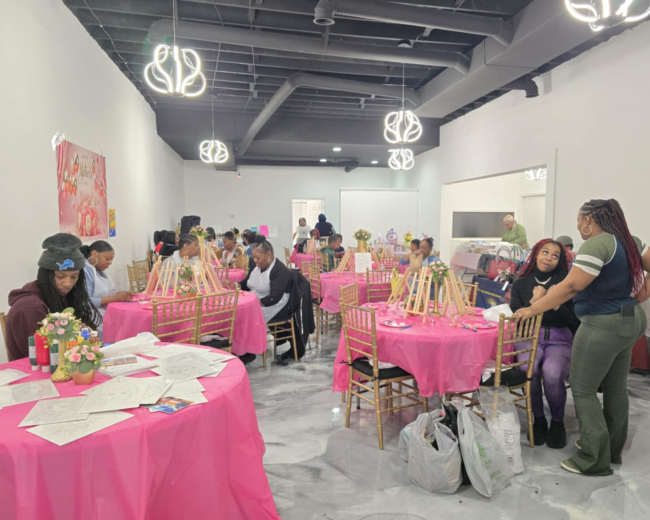 The image shows a vibrant event space filled with tables covered in pink cloth, where guests are engaged in an art activity, surrounded by floral arrangements and easels.