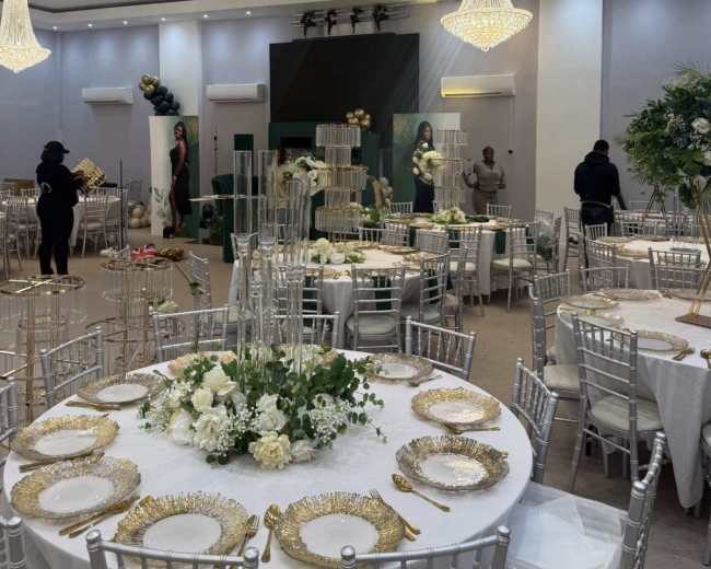 The image shows a spacious event hall set up for a formal occasion, featuring elegantly arranged tables with white tablecloths, floral centerpieces, and ornate chandeliers overhead.