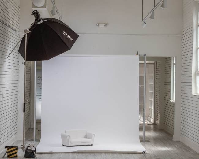 The image shows a minimalist photography studio with a white backdrop, a small white couch, and a large softbox light setup.
