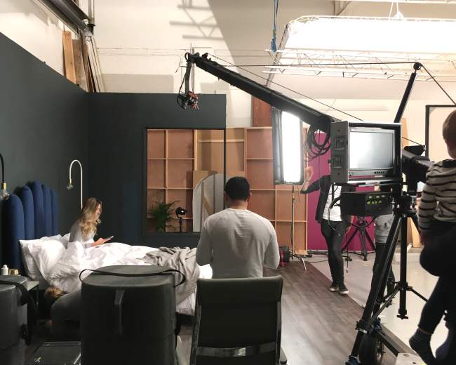 A film crew sets up a scene in a studio bedroom with a woman sitting on the bed and a camera mounted on a crane.