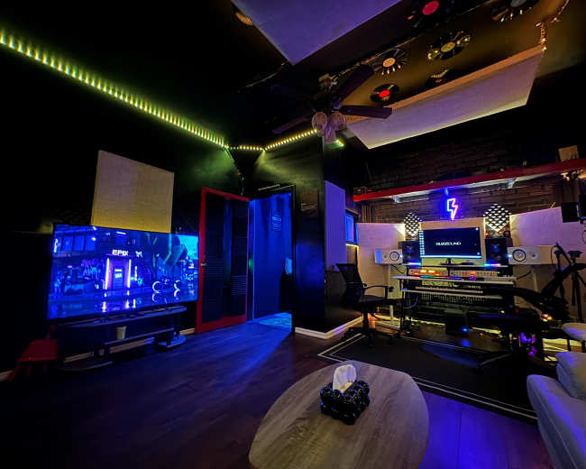 A dimly lit recording studio features a large screen, audio equipment, and mood lighting.