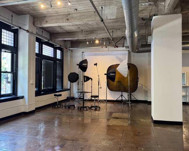 The image shows a photography studio equipped with lighting gear, softboxes, and a backdrop set up in a spacious room with large windows.