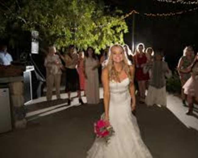 A bride in a white wedding dress stands in front of a group of women holding a bouquet, with string lights overhead creating a festive atmosphere.