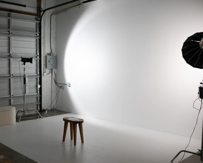 A photography studio with a wooden stool in front of a plain white backdrop, illuminated by studio lights.