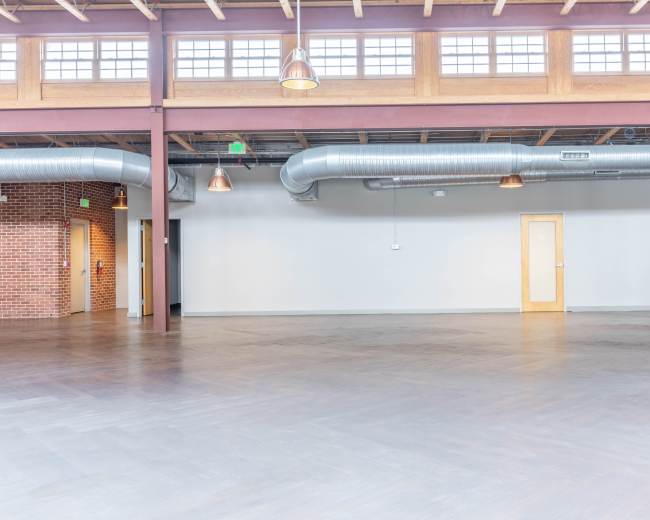 The image shows a spacious, industrial-style interior with exposed wooden beams, metal ductwork, and large windows.