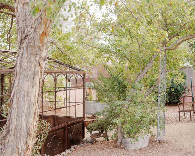 A metal greenhouse stands next to an arched entrance surrounded by trees and garden tools in a gravel area.