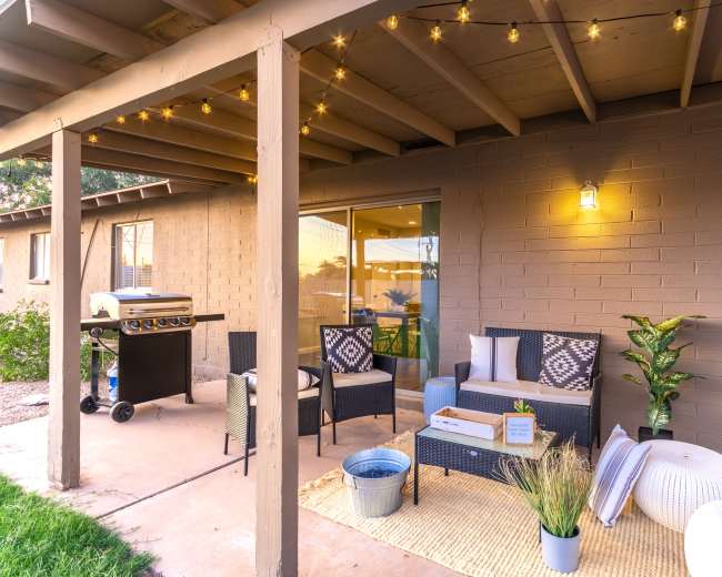 The image depicts a patio area featuring a seating arrangement with cushions, a grill, string lights overhead, and decorative plants.