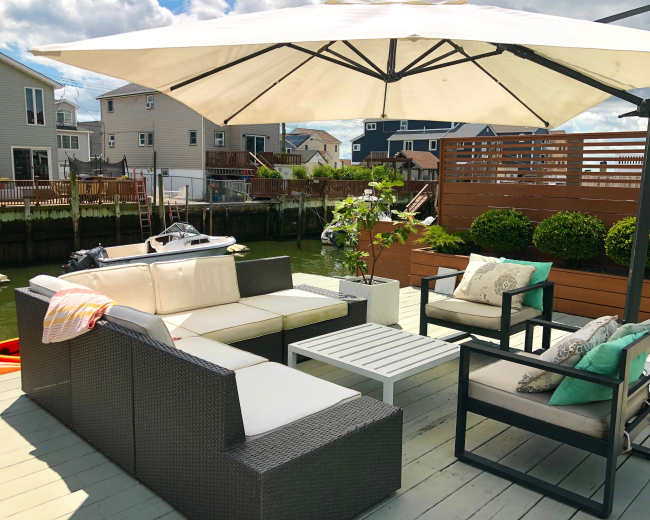 The image shows a patio area with a sectional sofa, armchairs, and a coffee table under a large umbrella, situated near a waterway and surrounded by houses.