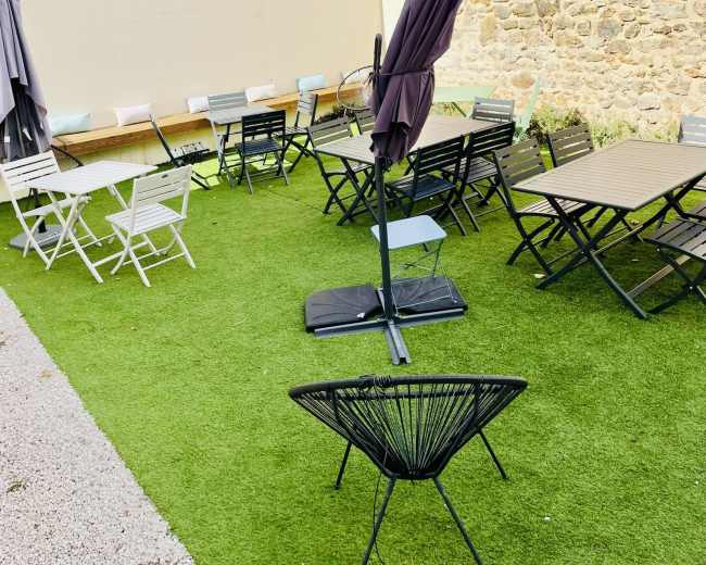 The image depicts an outdoor seating area featuring various tables and chairs on artificial grass, along with several umbrellas.