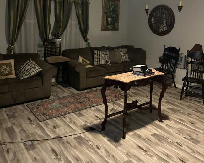 The image shows a living room with two couches, a coffee table, a wooden rocking chair, and decorative curtains, all arranged around a light-colored wooden floor.