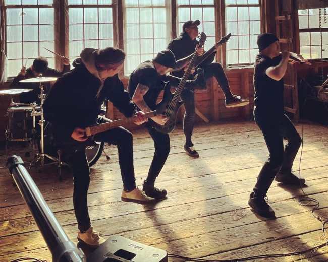 A band performs energetically in a rustic, sunlit room with wooden floors and large windows.