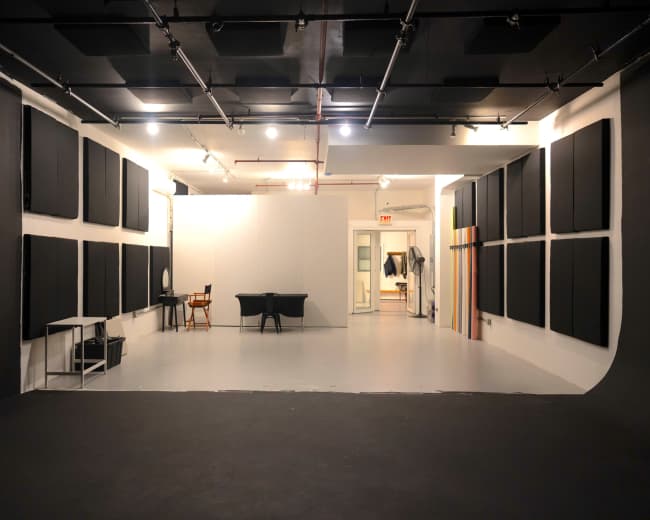 The image shows a spacious studio with black soundproofing panels on the walls, a large blank backdrop, and various pieces of furniture, including tables and chairs, in a well-lit environment.