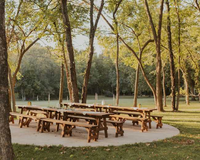 A circular wooden pavilion with several picnic tables is surrounded by tall trees in a grassy area.