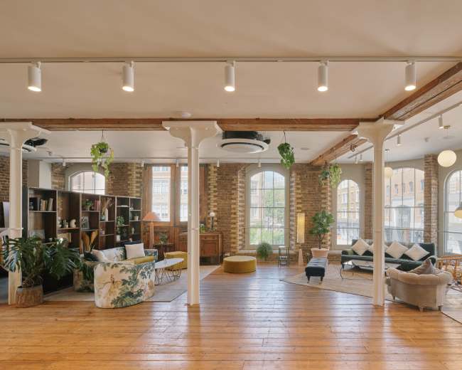 The image shows a spacious, well-lit interior featuring wooden floors, large windows, and a mix of seating areas with plants and decorative furniture.