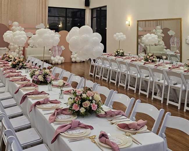 The image shows a elegantly arranged banquet hall with white tables set with pink napkins and floral centerpieces, featuring large chandeliers and decorative balloon displays.