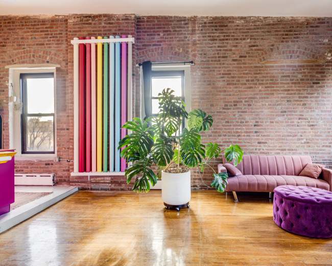 The image shows a modern living space with exposed brick walls, a purple tufted ottoman, a green potted plant, and a sofa facing large windows.