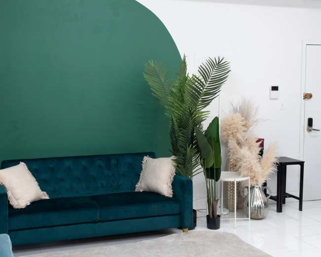 A dark green velvet sofa is positioned against a vibrant green curved wall, flanked by tall potted plants and a white side table with decorative items.