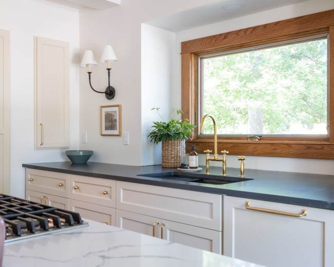 The image shows a modern kitchen with light-colored cabinetry, a black countertop, and a window framed in wood, featuring a sink and a potted plant nearby.