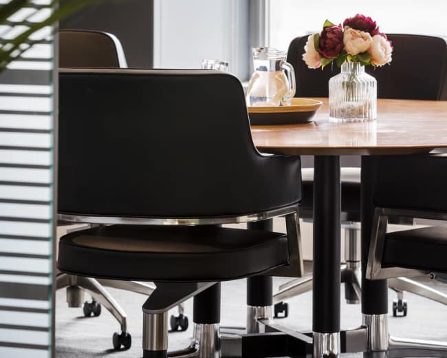 A modern meeting room features a wooden table surrounded by black ergonomic chairs, with a glass vase of flowers as a centerpiece.