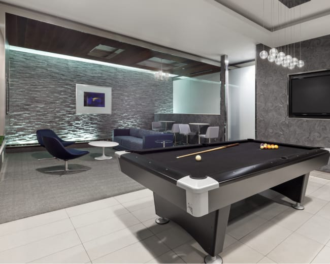 The image shows a modern game room featuring a black pool table, decorative wall panels, and a seating area with contemporary furniture.