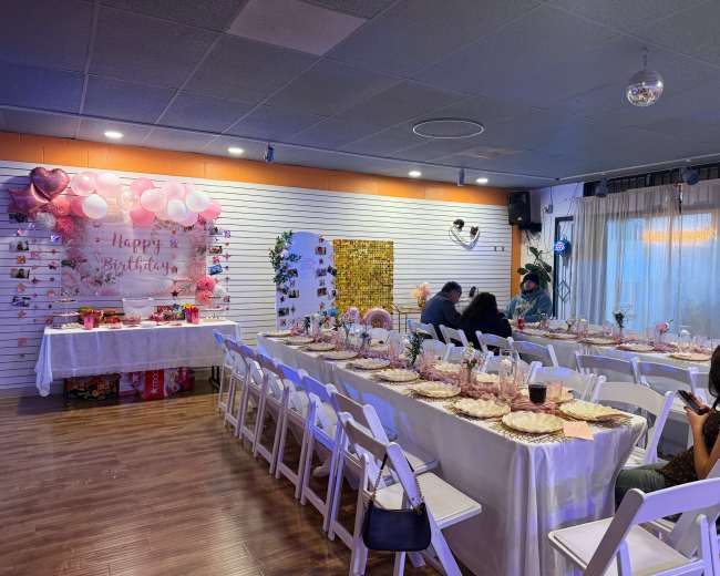 A decorated birthday party space features a long table set with white chairs, colorful decorations, and balloons along one wall.