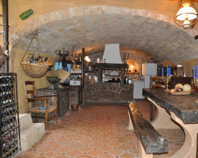 The image shows a rustic kitchen with stone walls, a large wooden table, and various items displayed on shelves and countertops.