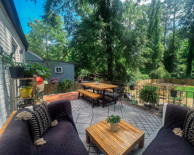 A patio area features wooden furniture surrounded by greenery and a fence in the background.