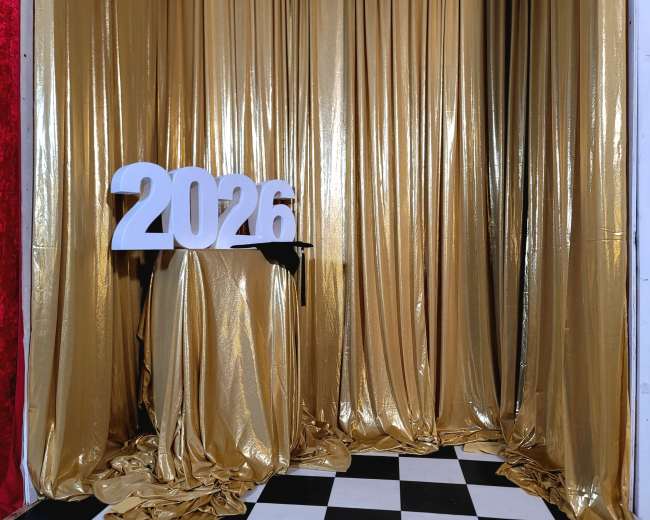 The image shows a gold curtain backdrop with the numbers "2026" displayed on a pedestal in front of a checkered floor.
