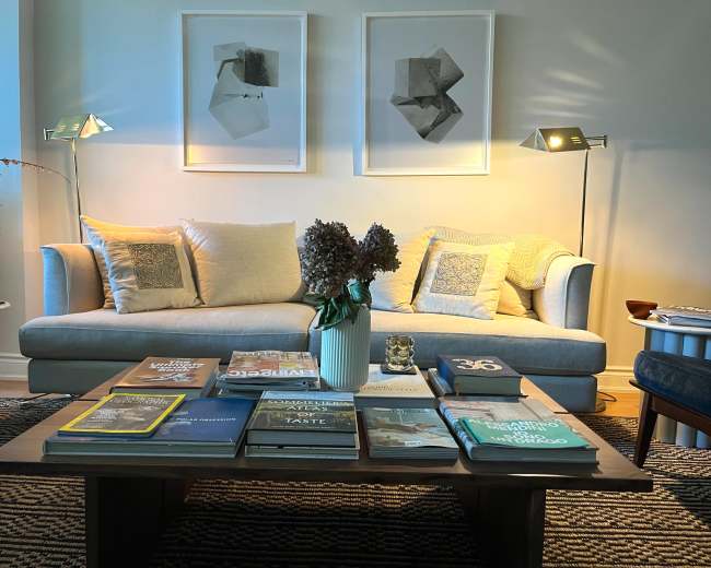 A modern living room features a light-colored sofa with decorative pillows, a wooden coffee table stacked with books, and two framed artworks on the wall, illuminated by stylish floor lamps.