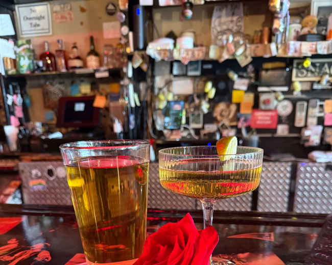 A glass of golden beer and a cocktail with a lime wedge sit next to a red rose on a bar countertop adorned with various decorations.