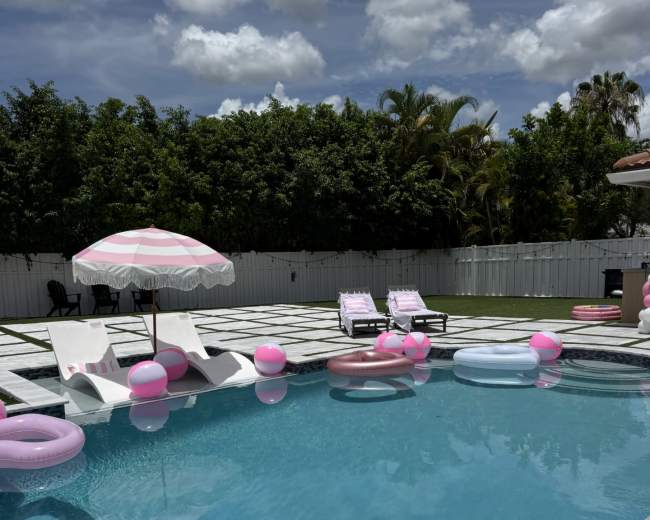 A backyard swimming pool features colorful inflatable floats, lounge chairs, and an umbrella under a partly cloudy sky.