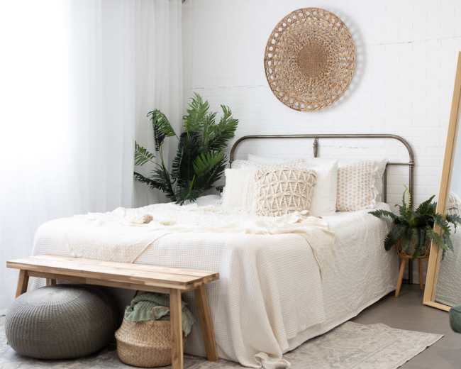 The scene shows a tidy bedroom with a bed dressed in neutral linens, a decorative wall hanging, potted plants, a large mirror, and a wooden bench at the foot of the bed.