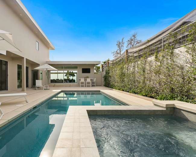 The image shows a modern outdoor pool area with a hot tub, lounge chairs, and umbrellas surrounded by a landscaped wall.
