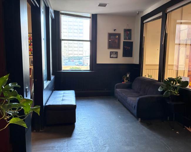 The image shows a small interior seating area featuring a dark couch, a bench, and large windows allowing natural light to enter.