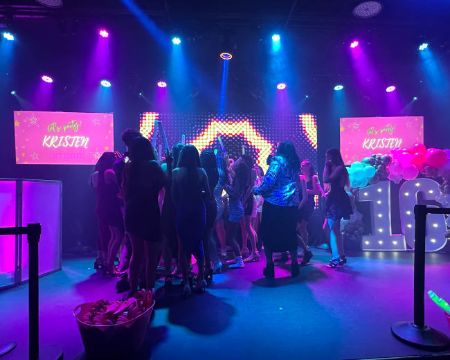 A group of young people is gathered on a stage celebrating a 16th birthday party, with colorful lights and decorations including a large "16" display.