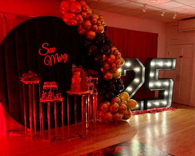 The image shows a decorated event space featuring a large illuminated "25" display, a backdrop with red and black balloons, and a table with desserts.