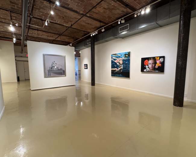 The image shows an art gallery with several framed artworks displayed on white walls and a polished floor under bright lighting.