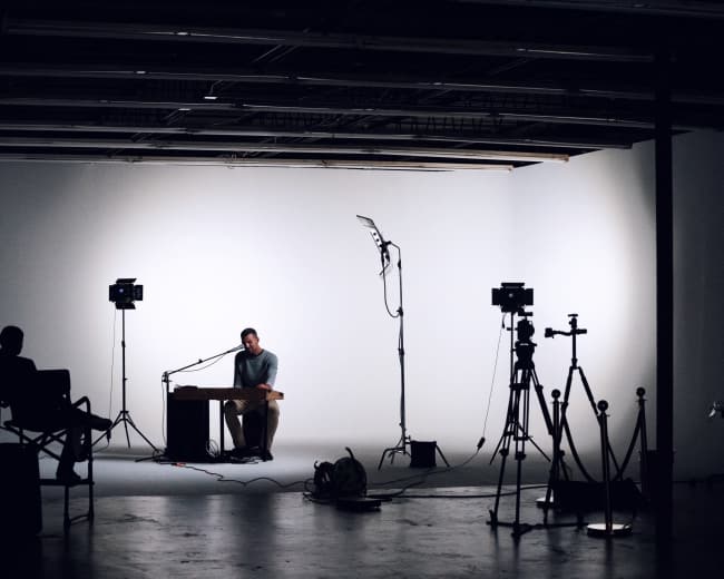 A musician sits at a table in a dimly lit studio with cameras and lighting equipment set up around him.